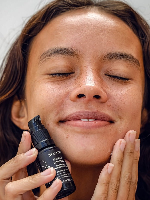 Woman with closed eyes holding Mukti Blemish Control Acne Spot Treatment 15ml beside her face while applying skincare.