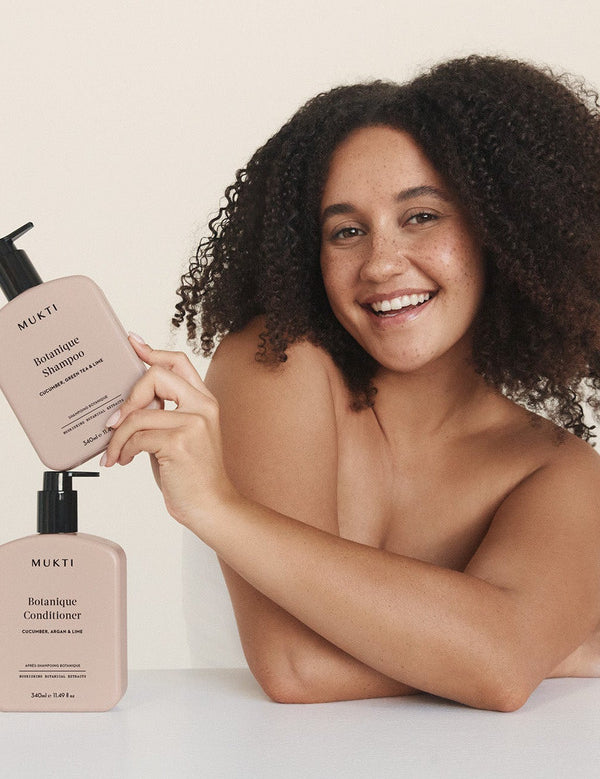 Smiling woman holding Mukti Botanique Conditioner above the matching bottle on a neutral studio surface.