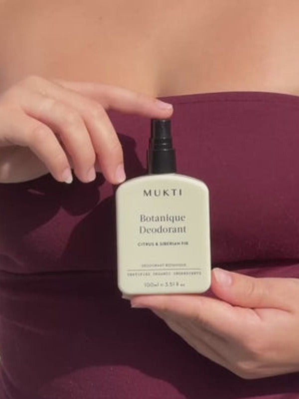 Person wearing a plum top holding Mukti Botanique Natural Deodorant Citrus & Siberian Fir against their torso.