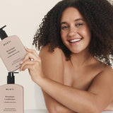 Smiling woman holding Mukti Botanique Shampoo above the matching bottle on a neutral studio surface.