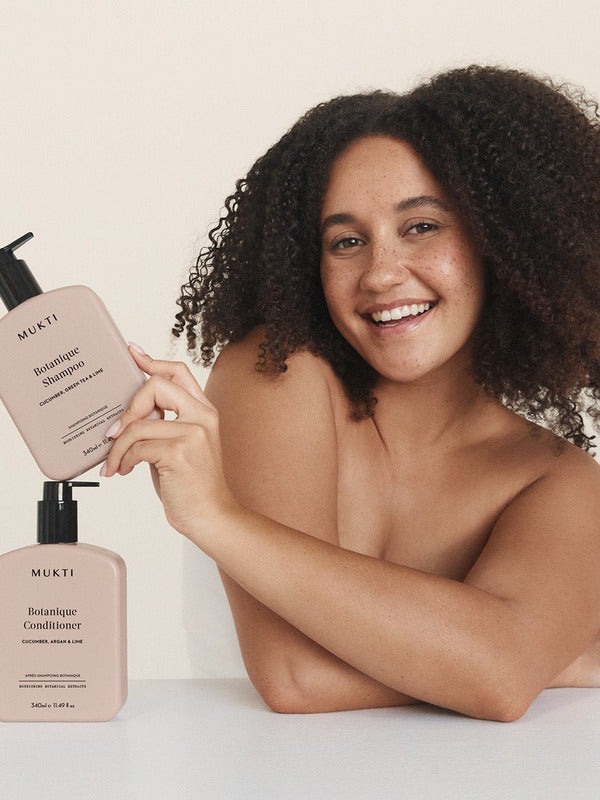 Smiling woman holding Mukti Botanique Shampoo above the matching bottle on a neutral studio surface.