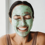 Smiling woman wearing a green layer of Mukti Deep Cleanse Enzyme Mask across her face.