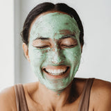 Smiling woman wearing a green layer of Mukti Deep Cleanse Enzyme Mask across her face.