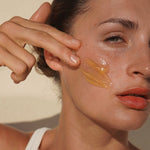 Woman massaging Mukti Gentle Melt Cleansing Balm onto her cheek beside the open jar.