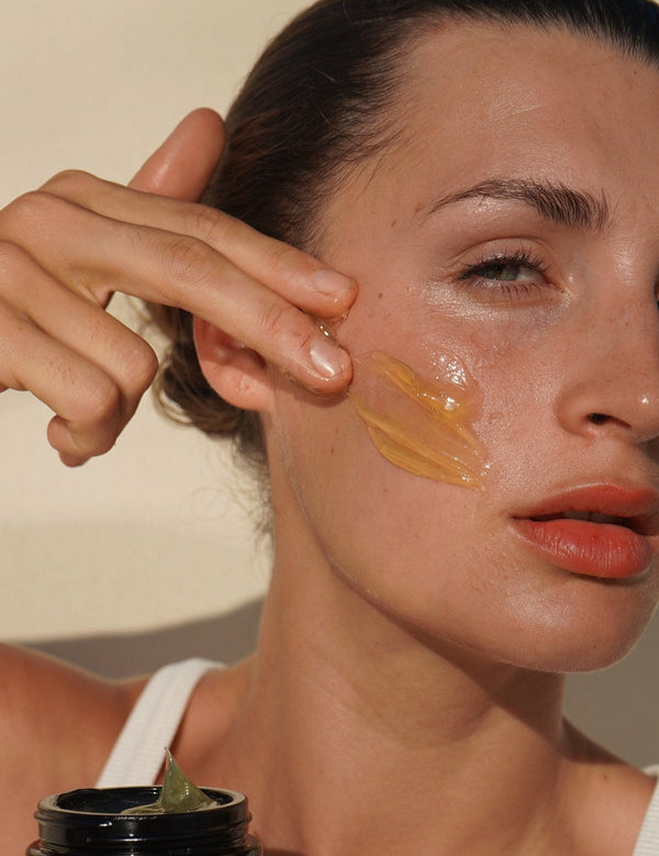 Woman massaging Mukti Gentle Melt Cleansing Balm onto her cheek beside the open jar.