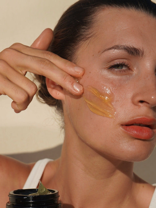 Woman massaging Mukti Gentle Melt Cleansing Balm onto her cheek beside the open jar.