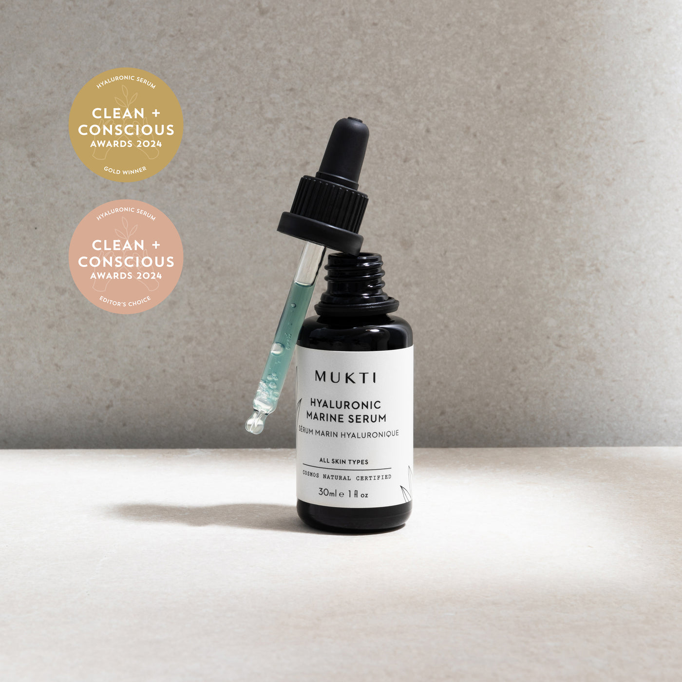 Open black Mukti Hyaluronic Marine Serum bottle with an aqua-filled dropper beside 2024 Clean and Conscious Gold Winner and Editor's Choice badges.