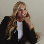 Blonde woman seated on a pale sofa with her chin resting on one hand, representing hydrated skin with Mukti Hydrating Moisturiser.