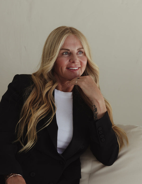 Blonde woman seated on a pale sofa with her chin resting on one hand, representing hydrated skin with Mukti Hydrating Moisturiser.
