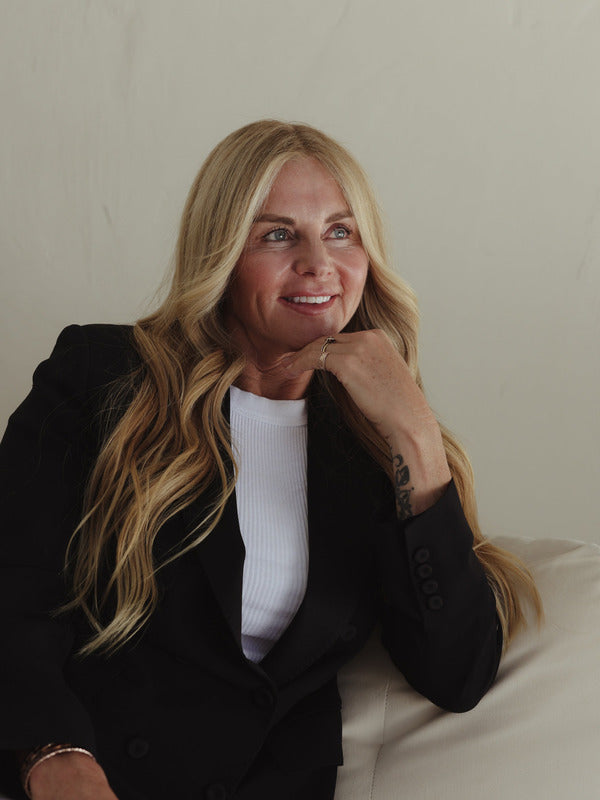 Blonde woman seated on a pale sofa with her chin resting on one hand, representing hydrated skin with Mukti Hydrating Moisturiser.