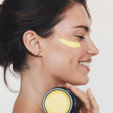Woman in profile holding an open jar of Mukti Marigold Hydrating Créme near her neck, with a golden cream swipe beneath her eye.