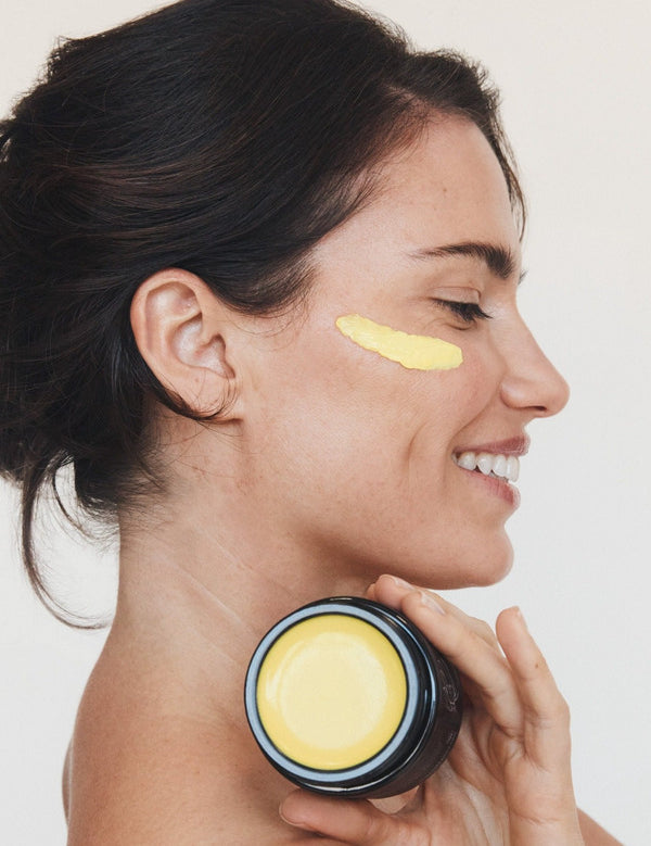Woman in profile holding an open jar of Mukti Marigold Hydrating Créme near her neck, with a golden cream swipe beneath her eye.