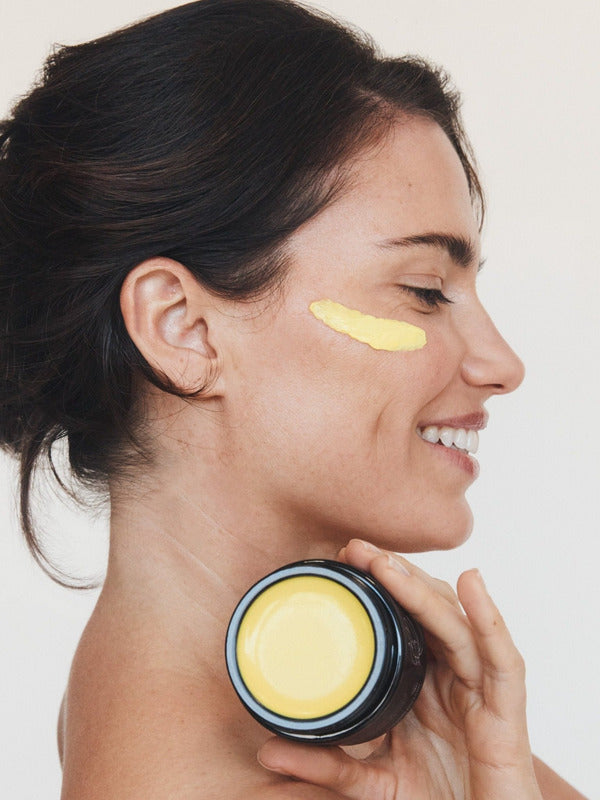 Woman in profile holding an open jar of Mukti Marigold Hydrating Créme near her neck, with a golden cream swipe beneath her eye.