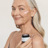 Silver-haired woman smiling while holding an open jar of Mukti Queen of the Night Crème and wearing the cream on her cheek.