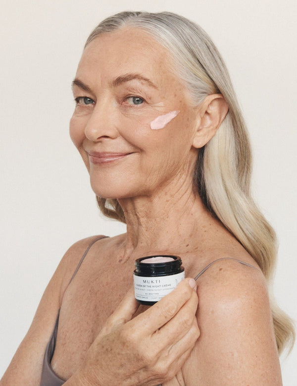 Silver-haired woman smiling while holding an open jar of Mukti Queen of the Night Crème and wearing the cream on her cheek.
