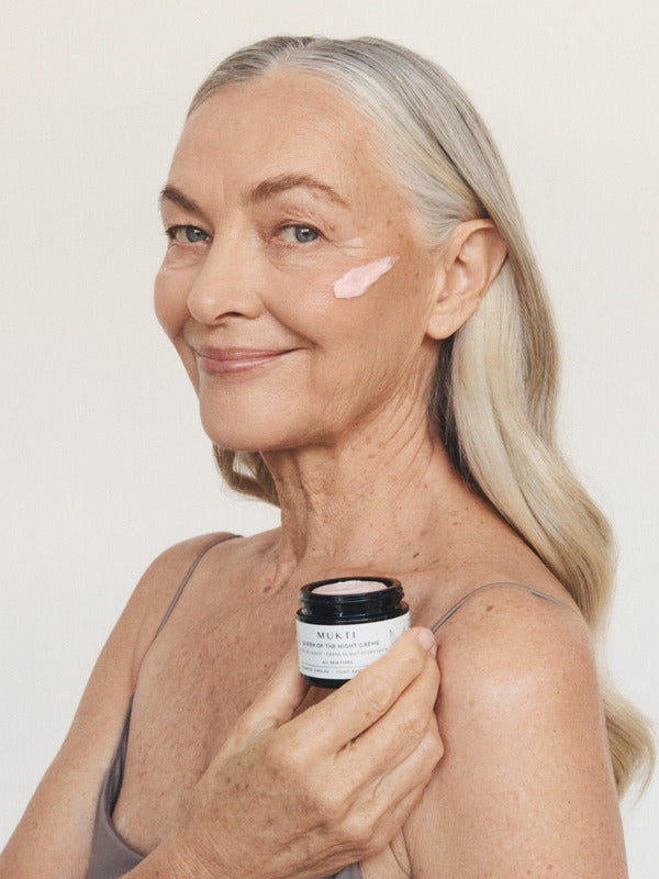 Silver-haired woman smiling while holding an open jar of Mukti Queen of the Night Crème and wearing the cream on her cheek.