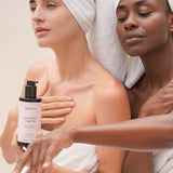 Two women in white towels applying Mukti Red Velvet Body Oil, with the bottle visible in the foreground.
