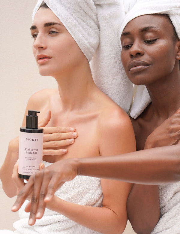 Two women in white towels applying Mukti Red Velvet Body Oil, with the bottle visible in the foreground.