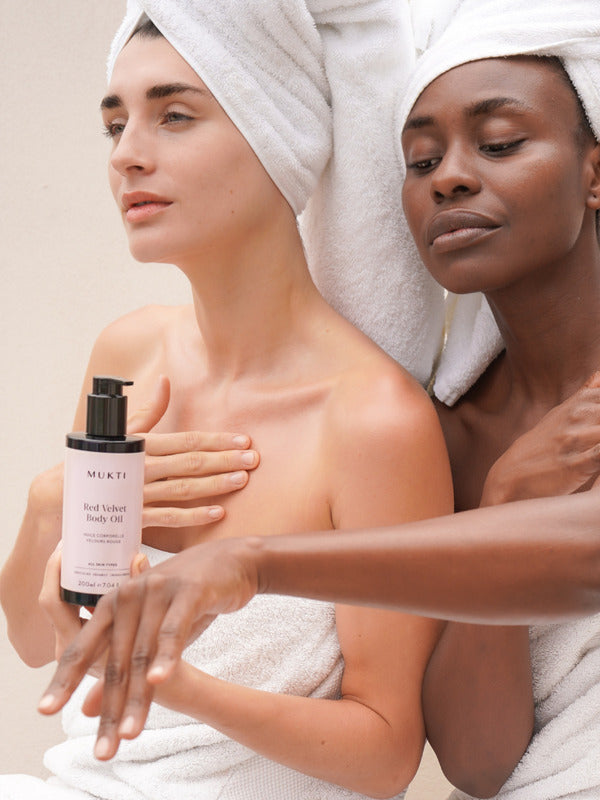 Two women in white towels applying Mukti Red Velvet Body Oil, with the bottle visible in the foreground.