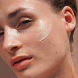 Close-up portrait of a woman with a curved swatch of Mukti Restorative Moisturiser applied to her cheek.