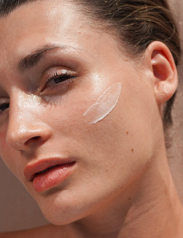 Close-up portrait of a woman with a curved swatch of Mukti Restorative Moisturiser applied to her cheek.