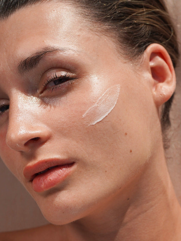 Close-up portrait of a woman with a curved swatch of Mukti Restorative Moisturiser applied to her cheek.