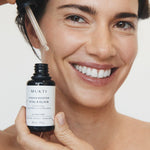 Woman smiling while holding Mukti Vitamin B Elixir and lifting serum from the bottle with the glass dropper.