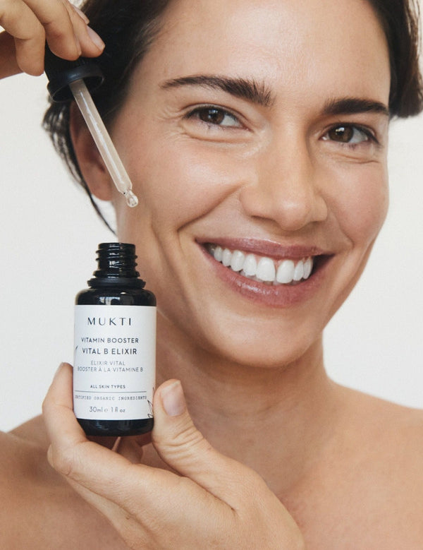 Woman smiling while holding Mukti Vitamin B Elixir and lifting serum from the bottle with the glass dropper.