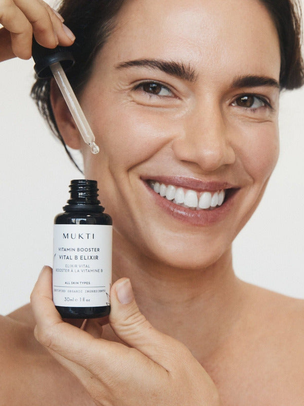 Woman smiling while holding Mukti Vitamin B Elixir and lifting serum from the bottle with the glass dropper.