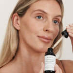 Blonde woman holding Mukti Vitamin C Elixir and glass dropper beside her face.