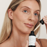 Blonde woman holding Mukti Vitamin C Elixir and glass dropper beside her face.
