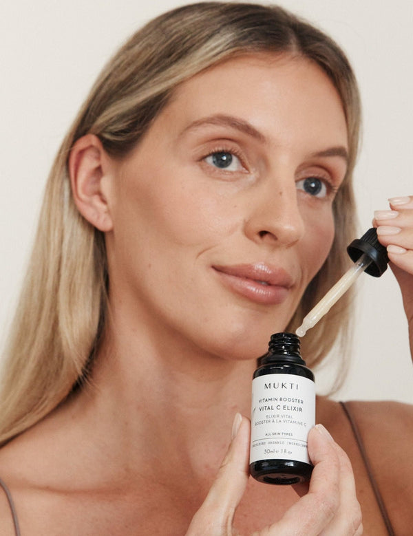 Blonde woman holding Mukti Vitamin C Elixir and glass dropper beside her face.