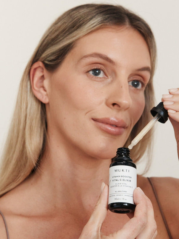 Blonde woman holding Mukti Vitamin C Elixir and glass dropper beside her face.