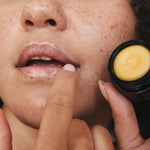 Close-up lifestyle photograph of a freckled woman applying Mukti Whipped Honey and Macadamia Lip Balm while holding the open black balm pot.
