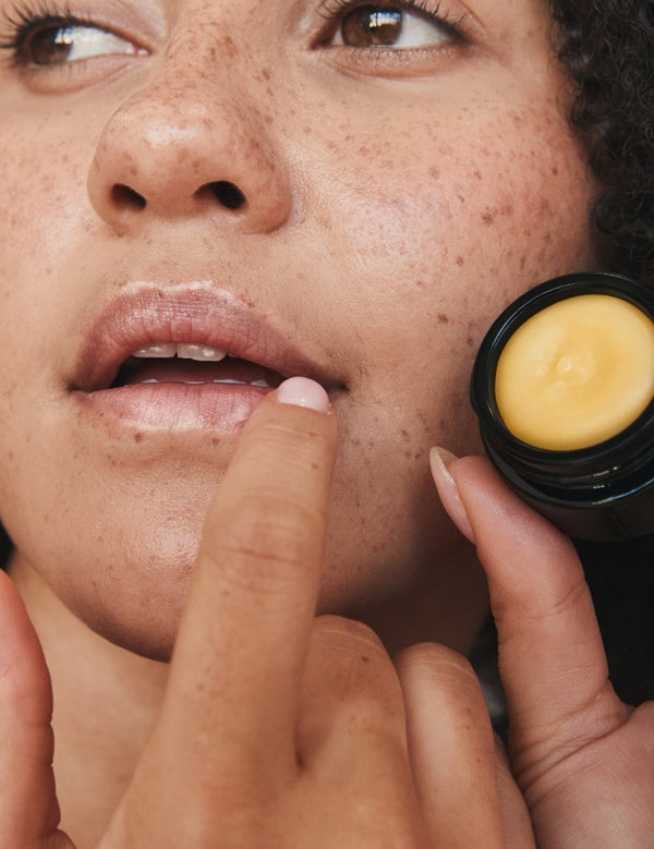 Close-up lifestyle photograph of a freckled woman applying Mukti Whipped Honey and Macadamia Lip Balm while holding the open black balm pot.
