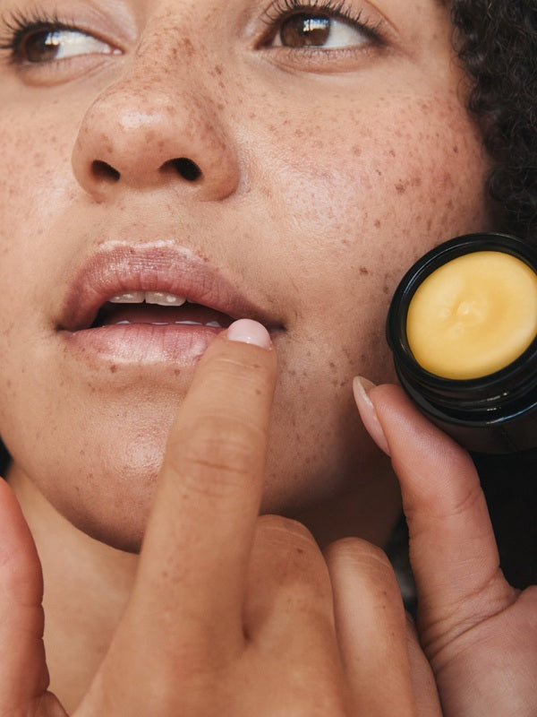 Close-up lifestyle photograph of a freckled woman applying Mukti Whipped Honey and Macadamia Lip Balm while holding the open black balm pot.