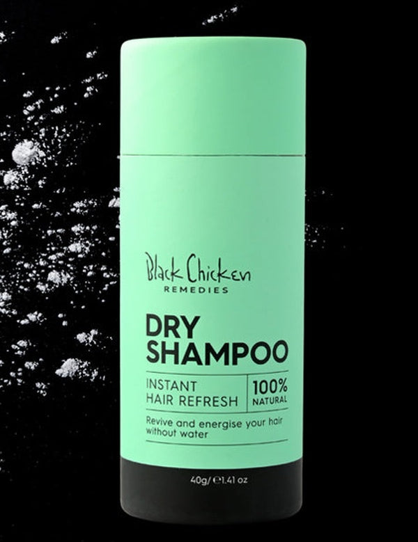 Mint-green Black Chicken Remedies Dry Shampoo tube with a black base, photographed beside scattered white powder on a black background.