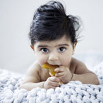 Baby lying on a chunky knit blanket while chewing a yellow Natural Rubber Fish Teether.