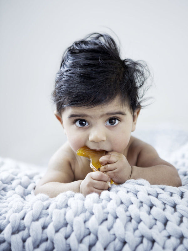 Baby lying on a chunky knit blanket while chewing a yellow Natural Rubber Fish Teether.