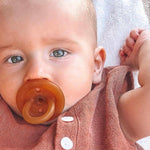 Close-up of a baby using an amber Natural Rubber Soother Small Orthodontic dummy while wearing a rust-coloured outfit.