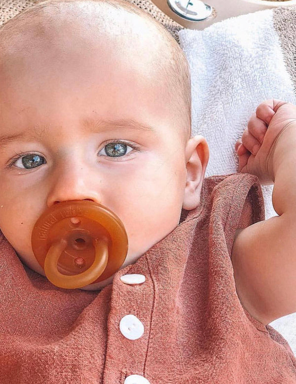 Close-up of a baby using an amber Natural Rubber Soother Small Orthodontic dummy while wearing a rust-coloured outfit.