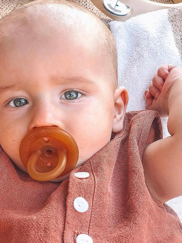 Close-up of a baby using an amber Natural Rubber Soother Small Orthodontic dummy while wearing a rust-coloured outfit.