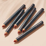 Six INIKA Organic Lip Crayons 6.7g, featuring pointed black barrels in assorted nude and red shades, are arranged diagonally on a beige surface, each casting a soft shadow.