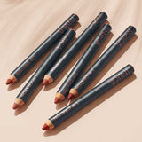 Six INIKA Organic Lip Crayons 6.7g, featuring pointed black barrels in assorted nude and red shades, are arranged diagonally on a beige surface, each casting a soft shadow.