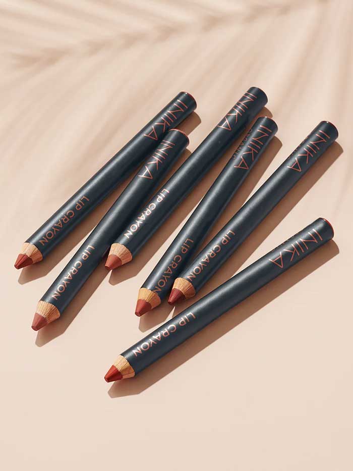Six INIKA Organic Lip Crayons 6.7g, featuring pointed black barrels in assorted nude and red shades, are arranged diagonally on a beige surface, each casting a soft shadow.