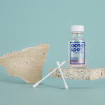 Noosa Basics Acne Spot Drying Lotion bottle styled with cotton buds and natural stone on a pale blue background.