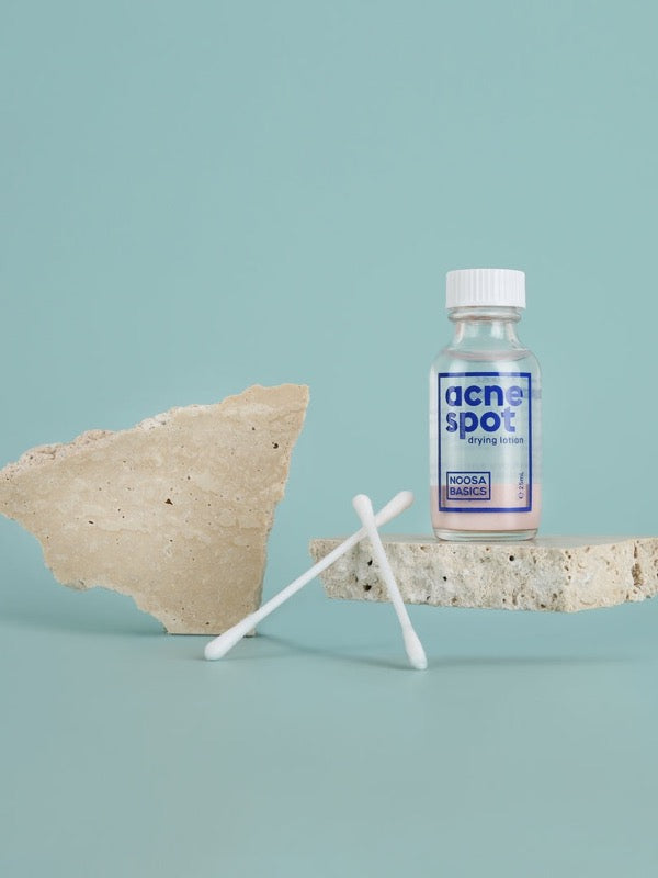 Noosa Basics Acne Spot Drying Lotion bottle styled with cotton buds and natural stone on a pale blue background.