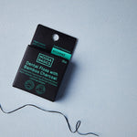 Black Noosa Basics Aniseed Bamboo Charcoal Dental Floss carton on a pale blue surface with a loose length of dark floss curling beside it.