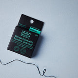 Black Noosa Basics Aniseed Bamboo Charcoal Dental Floss carton on a pale blue surface with a loose length of dark floss curling beside it.