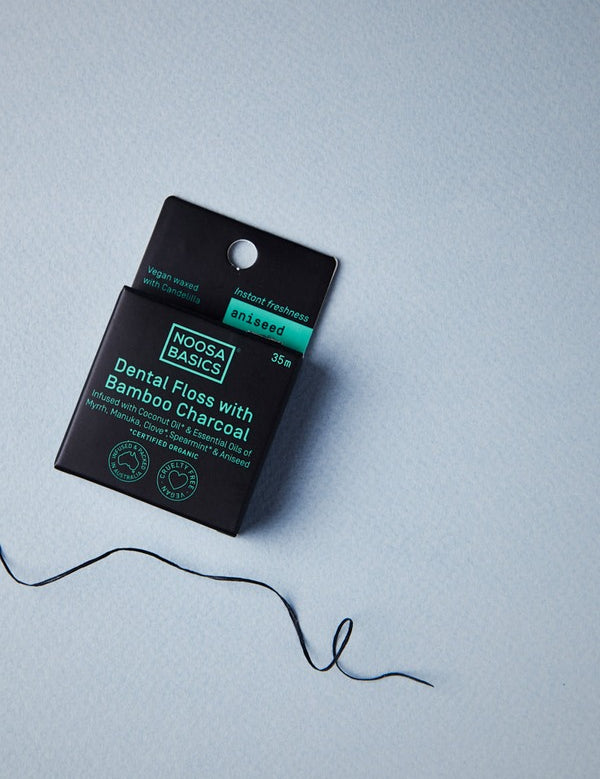 Black Noosa Basics Aniseed Bamboo Charcoal Dental Floss carton on a pale blue surface with a loose length of dark floss curling beside it.